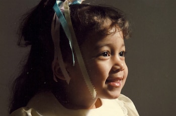 Retrato de perfil de una niña con cabello oscuro rizado, cintas de colores azul, rosa y blanco, y una prenda clara con encaje en el cuello. Fondo oscuro