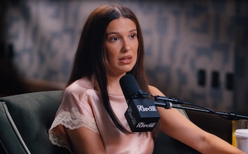 Millie Bobby Brown con blusa rosa de encaje sentada en silla verde oscuro. Un micrófono negro con el logo 'On Purpose' está frente a ella