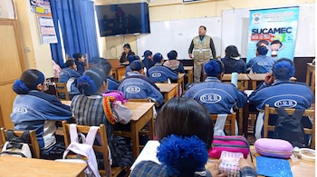 Un expositor habla frente a estudiantes sentados en carpetas dentro de un aula, junto a una pizarra blanca y un panel informativo