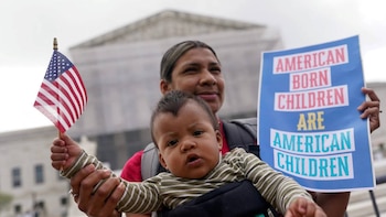 Mujer con bebé en portabebés. El bebé, vestido a rayas, sostiene una pequeña bandera estadounidense. La mujer sujeta un cartel azul con letras blancas y rojas