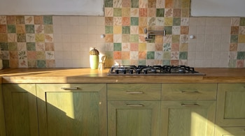 Cocina con gabinetes verdes, encimera de madera y estufa. Salpicadero de azulejos botánicos en tonos pastel, iluminado por el sol creando sombras.