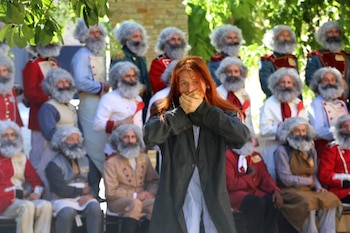 Persona con cabello rojo en primer plano cubre su boca con las manos. Detrás, un grupo de personas con pelucas rizadas, barbas postizas y uniformes de época