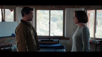 Andrew Rannells vistiendo chaqueta marrón y Allison Janney con suéter gris están frente a frente en una habitación