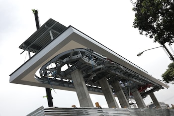 Vista desde abajo de la estructura de una estación de teleférico en construcción con pilares de hormigón, vigas metálicas y techo, junto a un árbol y una grúa
