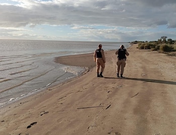 Dos efectivos de la Prefectura Naval Argentina caminan de espaldas por una playa de arena junto al agua