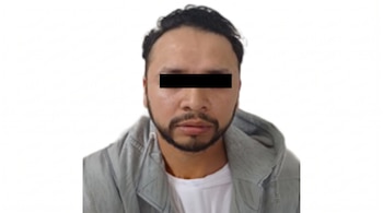 Hombre con barba y sudadera con capucha gris que tiene los ojos cubiertos por una franja negra sobre un fondo blanco.