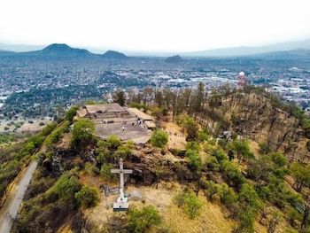 Vista aérea de cerro con plataforma de piedra, personas, gran cruz blanca, vegetación y camino. Al fondo, ciudad, montañas y torre de comunicaciones