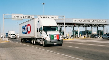 Un camión de carga blanco con la bandera de México y el logo del T-MEC en su tráiler cruza un carril exprés en una moderna frontera entre México y Estados Unidos, bajo cielo azul.