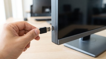 Una mano sostiene un conector USB negro frente al puerto lateral de un monitor de computadora sobre un escritorio.