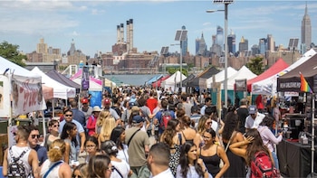 Una multitud de personas camina entre puestos de mercado al aire libre con toldos, con el East River y el horizonte de Manhattan al fondo en un día soleado