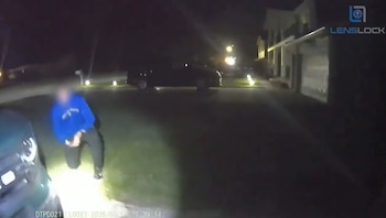Un hombre con sudadera azul está arrodillado en el césped por la noche, iluminado por una luz brillante. Un vehículo está en primer plano y otro en la calle