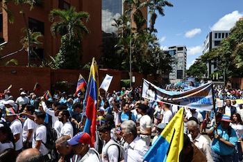 Multitud de personas marcha en una calle urbana con edificios y palmeras. Portan banderas de Venezuela y una pancarta con la imagen de María Corina Machado