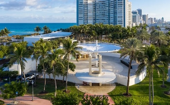 Vista aérea del Miami Beach Bandshell, un edificio blanco circular con un techo de platillo, rodeado de palmeras, frente al océano y edificios altos