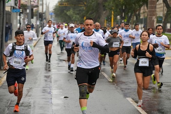 Corredores de diversas edades en una maratón urbana sobre calle mojada. Visten ropa deportiva, algunos con dorsales y edificaciones al fondo