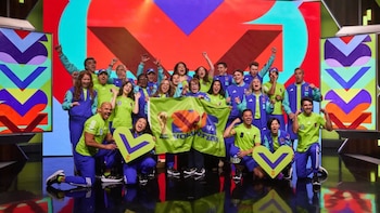 Un grupo numeroso de voluntarios sonríe y celebra, vestidos con uniformes verdes y azules. Sostienen la bandera 'FIFA Volunteer' y el trofeo de la Copa Mundial ante una pantalla LED colorida