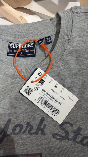 Etiqueta de cartón con código de barras y precios sujeta al cuello de una camiseta de color gris colgada de una percha de madera