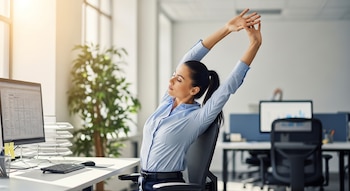 Mujer con camisa azul claro estirando los brazos hacia arriba mientras está sentada en una silla de oficina frente a un monitor en una oficina moderna y luminosa.