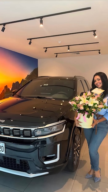 Mujer sonriente con cabello oscuro posa junto a camioneta Jeep negra, sosteniendo un ramo de flores en caja clara con lazo rosa. Fondo con mural de paisaje y techo con focos