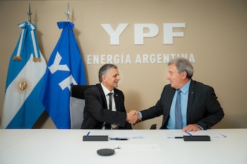 Dos hombres con traje se dan la mano en una mesa de reuniones. Detrás, la bandera argentina, la bandera de YPF y el logo "YPF Energía Argentina"