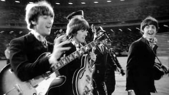 Fotografía en blanco y negro de John Lennon y Paul McCartney de The Beatles, caminando en un estadio con sus guitarras y trajes