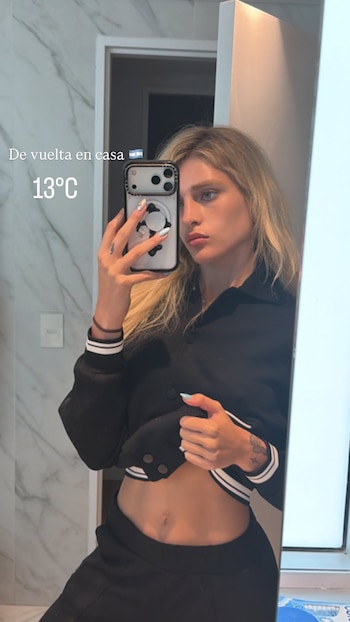 Mujer rubia frente a un espejo, tomando una selfie con un teléfono de funda blanca. Viste chaqueta negra y pantalón. Su abdomen está visible. Texto en pantalla: 'De vuelta en casa 🇦🇷 13°C'
