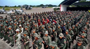 Un gran grupo de soldados uniformados con camuflaje y fusiles se forma en una pista aérea con aviones de combate y un helicóptero al fondo