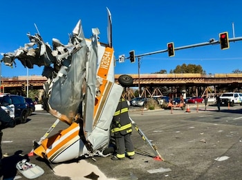 Una avioneta destruida en posición vertical sobre el pavimento, junto a un bombero, semáforos y vehículos de emergencia