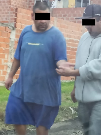 Dos hombres caminan frente a una pared de ladrillos con los ojos cubiertos por barras negras; uno viste de azul y el otro lleva chaqueta gris