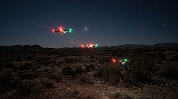 Tres drones con luces rojas y verdes vuelan de noche sobre un terreno desértico con vegetación baja y un cielo estrellado.