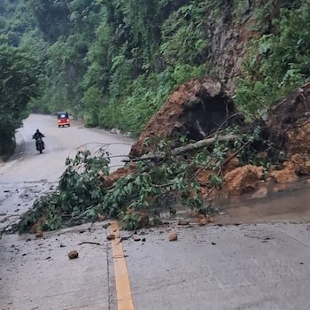 Rocas, tierra y ramas de árboles caídas sobre el asfalto de una carretera por la que transita un motociclista