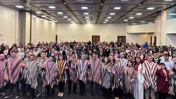 Un grupo numeroso de personas posa en un salón de eventos y levanta la mano, con varias autoridades en primera fila vistiendo ponchos andinos