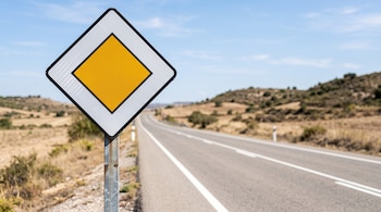Señal de tráfico en forma de rombo blanco con un cuadrado amarillo en el centro, situada junto a una carretera de asfalto.