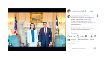 María Corina Machado, vistiendo un traje blanco y blusa azul, y Marco Rubio, con un traje oscuro y corbata, posan sonriendo en una sala formal con banderas