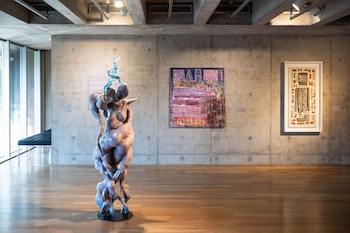 Una escultura abstracta y brillante de color lavanda y azul turquesa en el centro de una sala de museo con paredes de hormigón, suelo de madera y dos cuadros al fondo