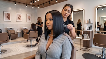Una estilista pasa un peine por el cabello negro y lacio de una mujer sentada en un sillón de peluquería.