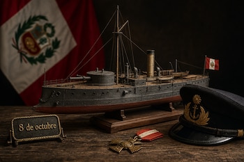 Maqueta del buque Huáscar sobre mesa de madera, con gorra naval, medalla, placa con la frase 8 de octubre y bandera peruana al fondo.