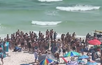 Vista aérea de una playa abarrotada con numerosas personas, algunas sombrillas de colores, olas en el mar y un vehículo con tablas de surf en la orilla