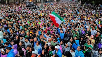 Gran multitud de personas en la calle, con vestimenta verde, blanca y roja, ponchos, y una bandera de México ondeando. Hay confeti blanco en el aire