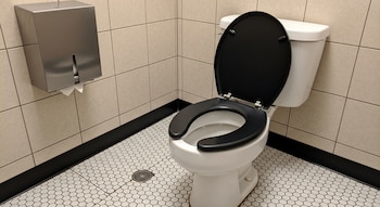 Un inodoro blanco con asiento y tapa negros abiertos en un baño público. Las paredes son de azulejos crema, el suelo de mosaico hexagonal y hay un dispensador metálico.