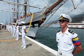 Oficial naval de Venezuela con uniforme blanco en primer plano. Fila de oficiales, buque de vela con personas, muelle, agua, montañas y otro barco al fondo