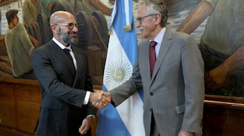 Dos hombres vestidos con traje se dan la mano. Detrás de ellos hay una bandera de Argentina y un mural. Uno usa lentes y tiene barba