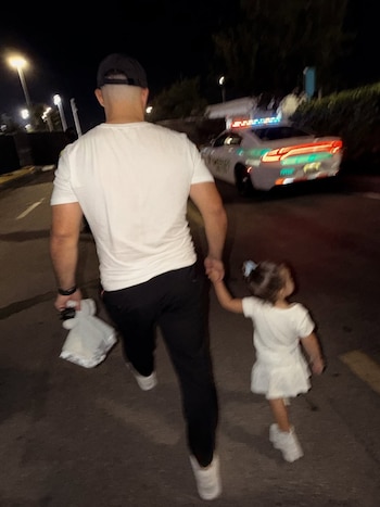 Hombre de espaldas con gorra y camiseta blanca, lleva una bolsa. Camina de la mano con una niña de vestido blanco. Al fondo, un coche policial con luces