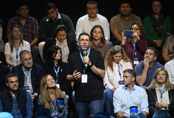 Hombre con barba y gafas habla en un micrófono en un evento político; a su alrededor, una audiencia diversa sentada lo observa
