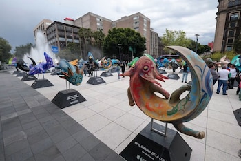 Numerosas esculturas coloridas con formas de criaturas fantásticas sobre pedestales negros en una plaza pavimentada con personas, edificios y una fuente