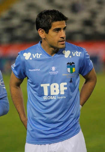 Hombre, futbolista Julio Barroso, de cabello oscuro, vestido con camiseta azul celeste de O'Higgins con lazo negro y pantalones blancos en el campo de juego
