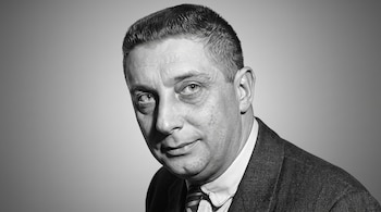 Plano medio de un hombre en blanco y negro que viste saco, camisa y corbata a rayas frente a un fondo claro.