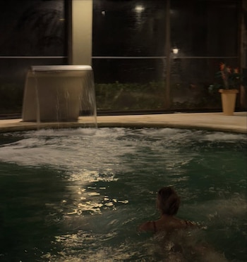 Vista trasera de una persona en una piscina climatizada con cascada de agua, burbujas y reflejos de luz. Ventanales oscuros de fondo y una planta en maceta