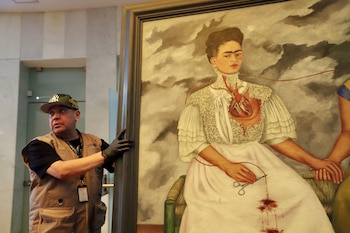 Hombre con gorra, chaleco y guantes negros mueve un gran cuadro enmarcado que representa a una mujer con un corazón expuesto y tijeras