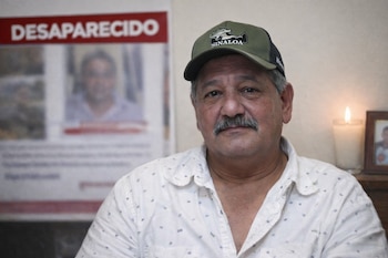 Hombre de mediana edad con bigote, gorra verde con 'Sinaloa' y camisa clara, posando frente a un cartel borroso de 'Desaparecido' y una vela encendida.