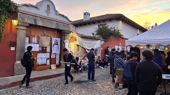 Personas se reúnen en una calle adoquinada con edificios históricos y pancartas; una carpa blanca y un cielo de atardecer son visibles